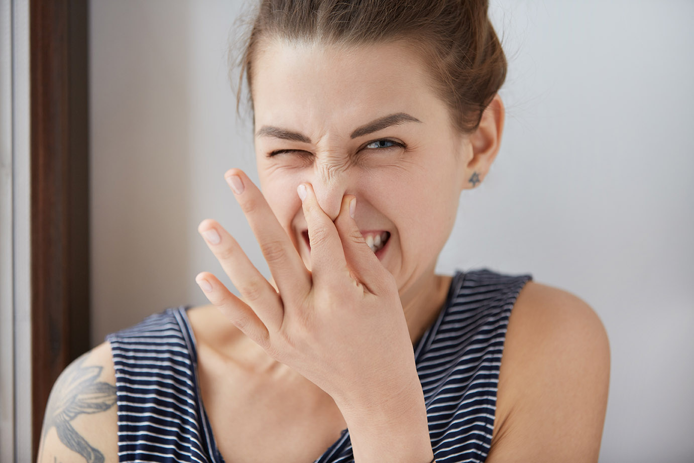 Close-up shot of Caucasian girl showing disgust, pinching her nose to avoid bad smell. Brunette girl with bunch of hair narrowing eyes in aversion to awful stink. Negative emotions, nasty feelings.